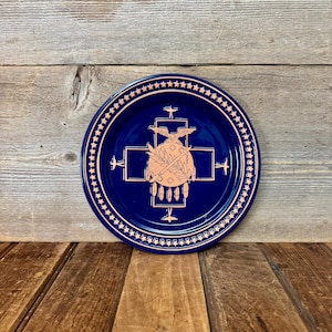 May include: A circular dark blue decorative plate featuring a copper-colored design. The border is lined with a ring of stars. The center displays a cross shape, an eagle, a circular emblem with a sword and olive branch, and hanging feathers.