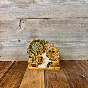 May include: A ceramic figurine featuring a spinning wheel, a dog, and a cat. The figurine is primarily brown and gold, with a white dog. The design is detailed and has a vintage aesthetic.