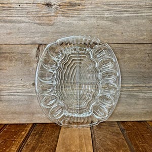 May include: Clear glass egg serving platter with oval egg-shaped indentations around the edge and a central section. The platter is oval-shaped with a scalloped edge. The background is a wooden surface.