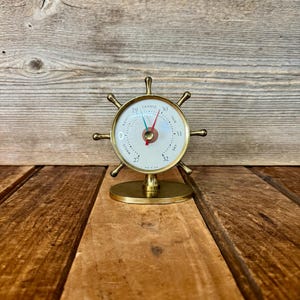 May include: A brass ship wheel barometer with a circular face. The face has a needle pointing to the current atmospheric pressure, with markings for "Storm", "Fair", and "Change". The barometer sits on a round base.