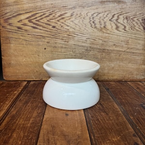 May include: A white ceramic butter dish with a rounded base and a shallow, circular top. The dish has a smooth, glossy finish and is designed to hold butter. The background is a wooden surface.