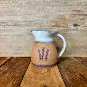 May include: A ceramic pitcher with a white top and handle, a light blue rim, and a brown body. The body features an embossed wheat design. The pitcher is displayed on a wooden surface.