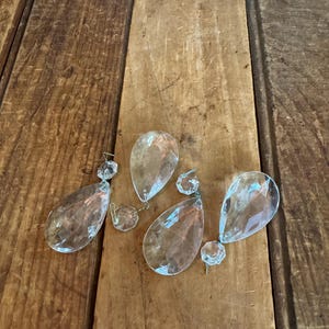 May include: Clear crystal chandelier pendants in a teardrop shape, with smaller round crystals attached. The pendants are displayed on a rustic, wooden surface, showcasing their reflective qualities.