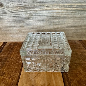 May include: A clear, rectangular glass box with a lid. The box features a geometric pattern and the letter "A" on the lid. The box is sitting on a wooden surface.