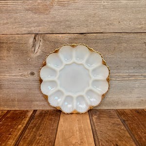 May include: A white, scalloped-edge serving dish with a gold rim. The dish has a central circular area surrounded by 12 petal-shaped compartments. The dish is made of opaque glass and sits on a wooden surface.