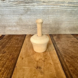 May include: A small, unfinished wooden kitchen tool with a rounded top and a cylindrical handle. The tool has a bulbous base and is set against a rustic wooden background. This item is likely used for food preparation.