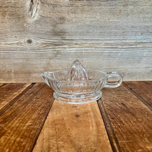 May include: Clear glass citrus juicer with a ribbed bowl, a pointed reamer, and a small handle. The juicer sits on a round base. The background is a wooden surface.