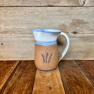 May include: A handmade ceramic pitcher with a wheat stalk design. The pitcher features a brown base, a light blue band, and a white top with a blue rim. The handle is white. The pitcher is displayed on a wooden surface.