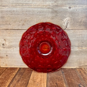 May include: A round, red glass bowl with a textured surface and a gold rim. The bowl has a scalloped edge and sits on a wooden surface. The bowl is a decorative piece.