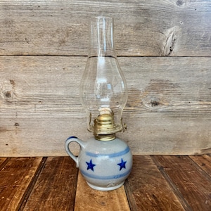 May include: A vintage oil lamp with a clear glass chimney and a gray ceramic base. The base features blue stripes and star designs. The lamp has a brass burner and a handle. The lamp is approximately 8 inches tall.