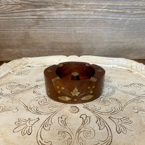 May include: A small, round wooden ashtray with brass inlay. The ashtray has a floral design and is sitting on a white tray with a floral pattern.