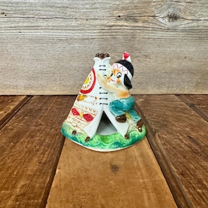 May include: A vintage ceramic figurine depicting a Native American child in traditional attire, leaning against a white teepee. The teepee is decorated with colorful patterns and sits on a green base. The figurine is on a wooden surface.