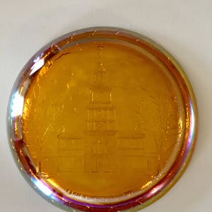 Peut inclure: Une assiette ronde en verre ambr&eacute; avec un motif grav&eacute; d'un b&acirc;timent avec un clocher. L'assiette a un bord iridescent, semblable &agrave; un arc-en-ciel.