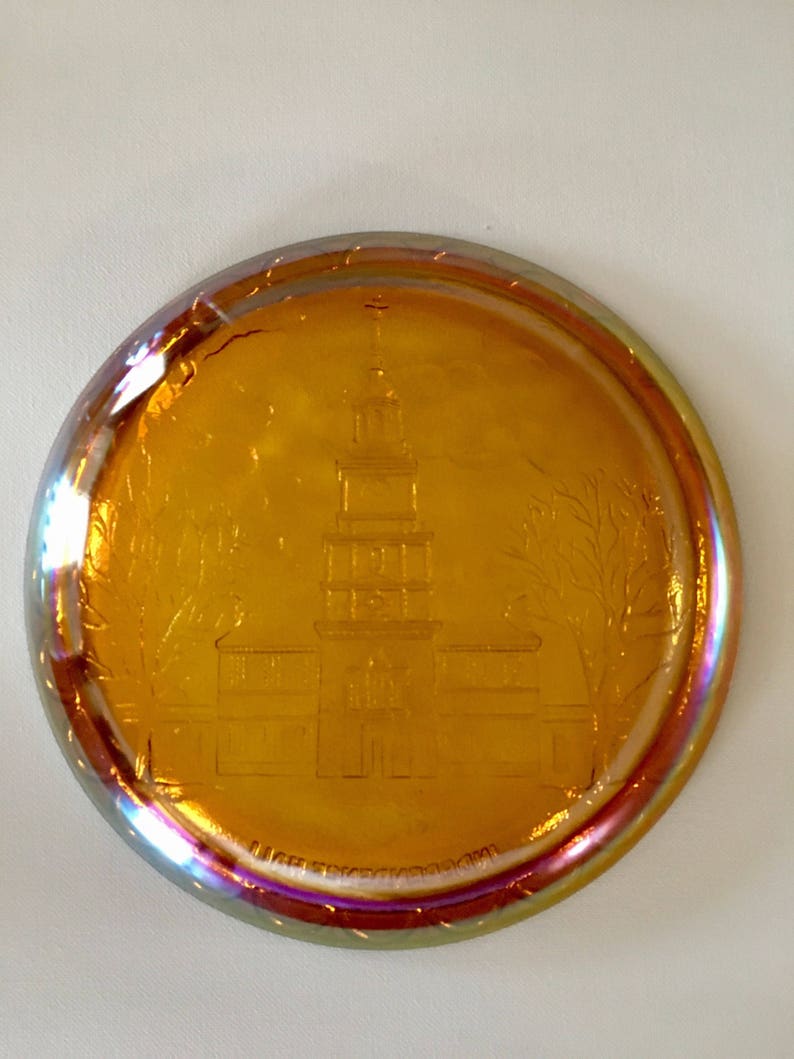 Peut inclure: Une assiette ronde en verre ambr&eacute; avec un motif grav&eacute; d'un b&acirc;timent avec un clocher. L'assiette a un bord iridescent, semblable &agrave; un arc-en-ciel.