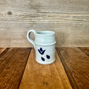 May include: A white ceramic mug with a black handle and the text "But first, coffee" in a black, handwritten-style font. The mug is on a white surface.