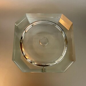 May include: A clear glass ashtray with an octagonal shape and a round center. The ashtray has a small hole in the center.