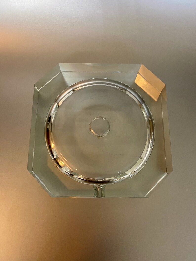 May include: A clear glass ashtray with an octagonal shape and a round center. The ashtray has a small hole in the center.