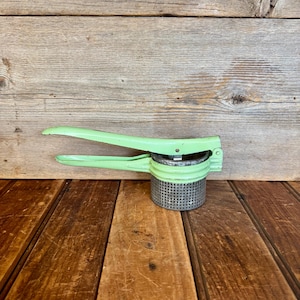May include: A vintage green metal potato masher with a handle and a perforated metal base. The masher is sitting on a wooden surface.
