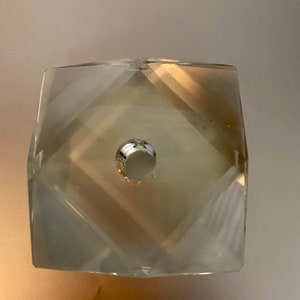 May include: A clear glass crystal prism with a faceted design and a hole in the center. The prism is a light beige color.