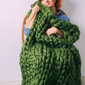 May include: A woman sits in a chair wrapped in a large, chunky knit green blanket. The blanket is made of thick yarn and has a loose, open weave.