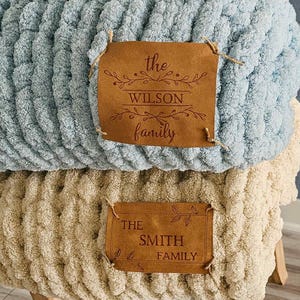 May include: Two chunky knit blankets, one light blue and one beige, stacked on a wooden stool. Each blanket has a brown leather tag with family names engraved. The top blanket reads "The Wilson Family".