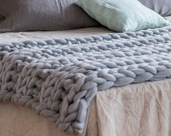 Puede incluir: Una cabecera acolchada de terciopelo rosa polvoriento con una falda de cama de lino blanco y una manta de punto grueso de color gris. La cama está hecha con almohadas grises y blancas y una almohada de tiro verde claro.