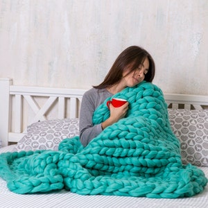 May include: A woman is sitting on a bed wrapped in a large, chunky knit teal blanket. She is holding a red mug in her hands.