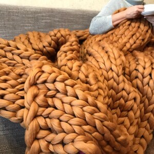 Op de afbeelding: Een groot, grof gebreid deken in een warme bruine kleur. Het deken is gemaakt van dik garen en heeft een zachte, knusse textuur.