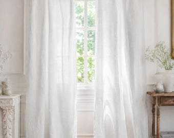 May include: Two white linen curtains hang from a wooden rod, tied with light-colored ribbons. The curtains frame a window with a view of green foliage. The room has white walls and a rustic wooden table.