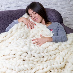 May include: A woman is curled up on a couch with a large, white chunky knit blanket. She is smiling and appears to be sleeping.