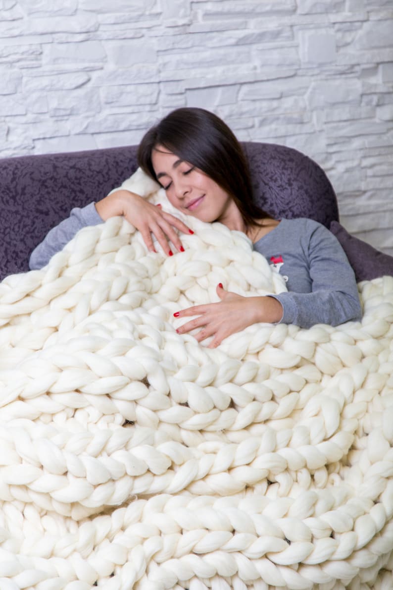 May include: A woman is curled up on a couch with a large, white chunky knit blanket. She is smiling and appears to be sleeping.