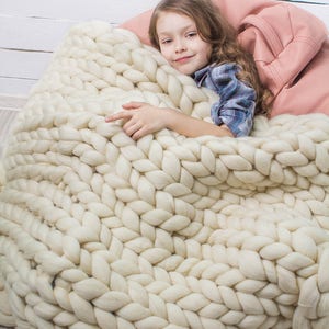 May include: A young girl is curled up in a large, white, chunky knit blanket. The blanket is made of thick, soft yarn and has a cable knit pattern. The girl is smiling and looks comfortable.