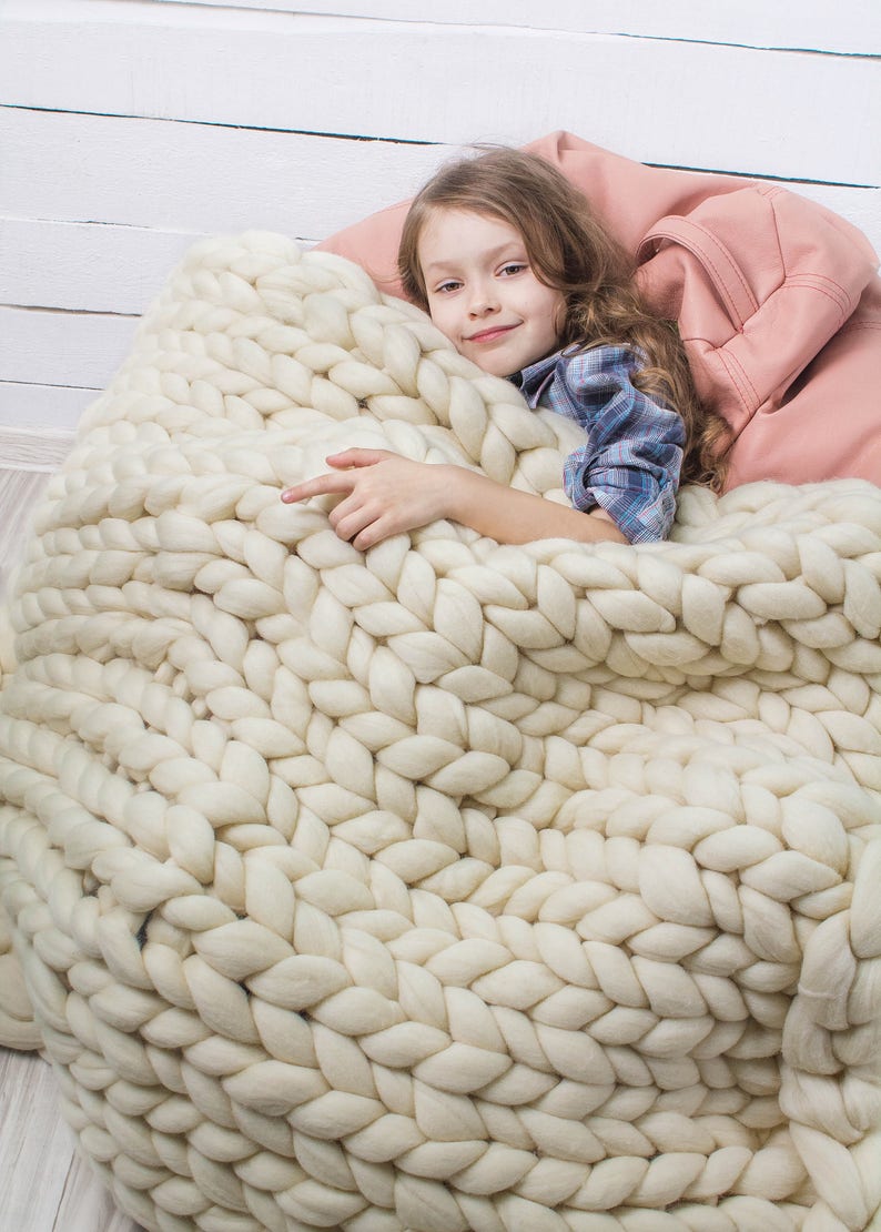 May include: A young girl is curled up in a large, white, chunky knit blanket. The blanket is made of thick, soft yarn and has a cable knit pattern. The girl is smiling and looks comfortable.