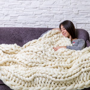 May include: A woman is relaxing on a couch, wrapped in a large, chunky knit white blanket. The blanket is made of thick, soft yarn and has a cozy, comforting look.