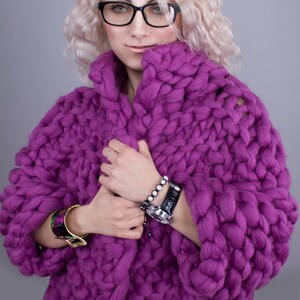 K&ouml;nnte beinhalten: Ein klobiger, &uuml;bergro&szlig;er lila Strickcardigan. Der Pullover hat einen leuchtenden Lilaton und ein dickes, strukturiertes Aussehen. Das Model tr&auml;gt eine Brille mit schwarzem Rahmen und ein Armband. Der Cardigan scheint aus einem weichen, warmen Material zu sein.