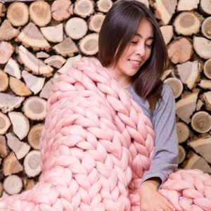 May include: A woman is wrapped in a large, chunky knit pink blanket. The blanket is made of thick yarn and has a soft, cozy texture. The woman is sitting in front of a wall of stacked logs.