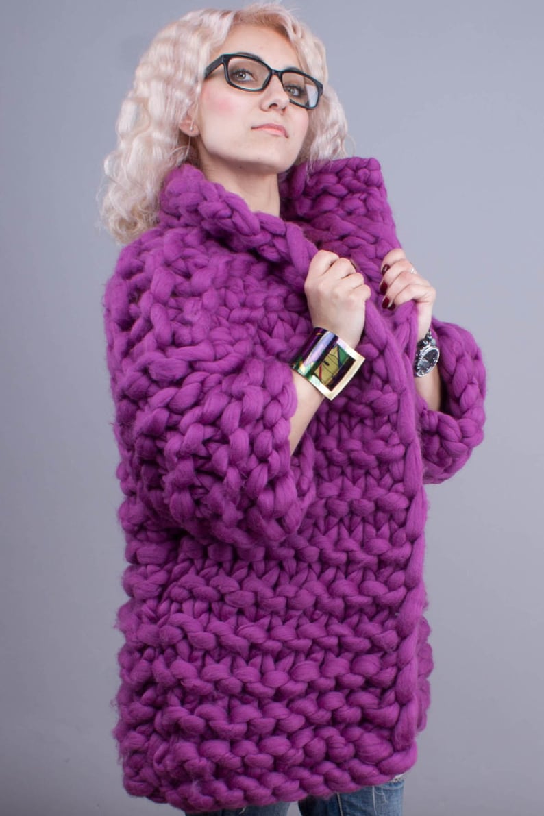 K&ouml;nnte beinhalten: Ein klobiger, handgestrickter, &uuml;bergro&szlig;er lila Pullover mit hohem Kragen. Der Pullover ist aus dickem Garn gefertigt und hat ein strukturiertes Aussehen. Die &Auml;rmel sind lang und der Stil ist gem&uuml;tlich und warm. Ein farbenfrohes Armband und eine Uhr sind sichtbar.