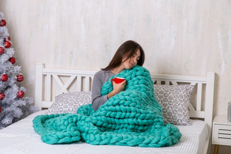 May include: A woman is sitting on a bed wrapped in a large, chunky knit teal blanket. She is holding a red mug in her hands.