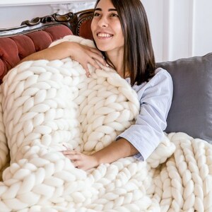 May include: A woman is sitting on a couch and is wrapped in a white chunky knit blanket. The blanket is made of thick yarn and has a large, loose knit stitch.