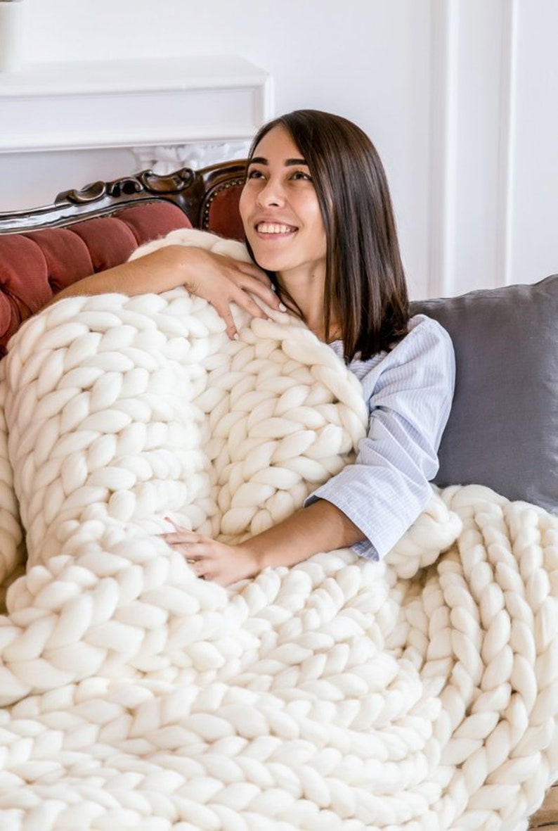 May include: A woman is sitting on a couch and is wrapped in a white chunky knit blanket. The blanket is made of thick yarn and has a large, loose knit stitch.