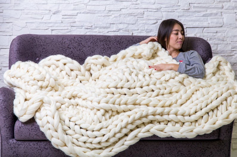 May include: A white chunky knit blanket draped over a purple couch. A person is sitting on the couch, partially covered by the blanket.