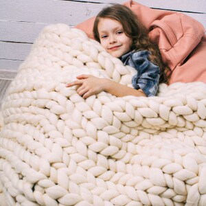 May include: A young person is curled up in a large, white, chunky knit blanket. The blanket is made of thick, soft yarn and has a cozy, comforting look.