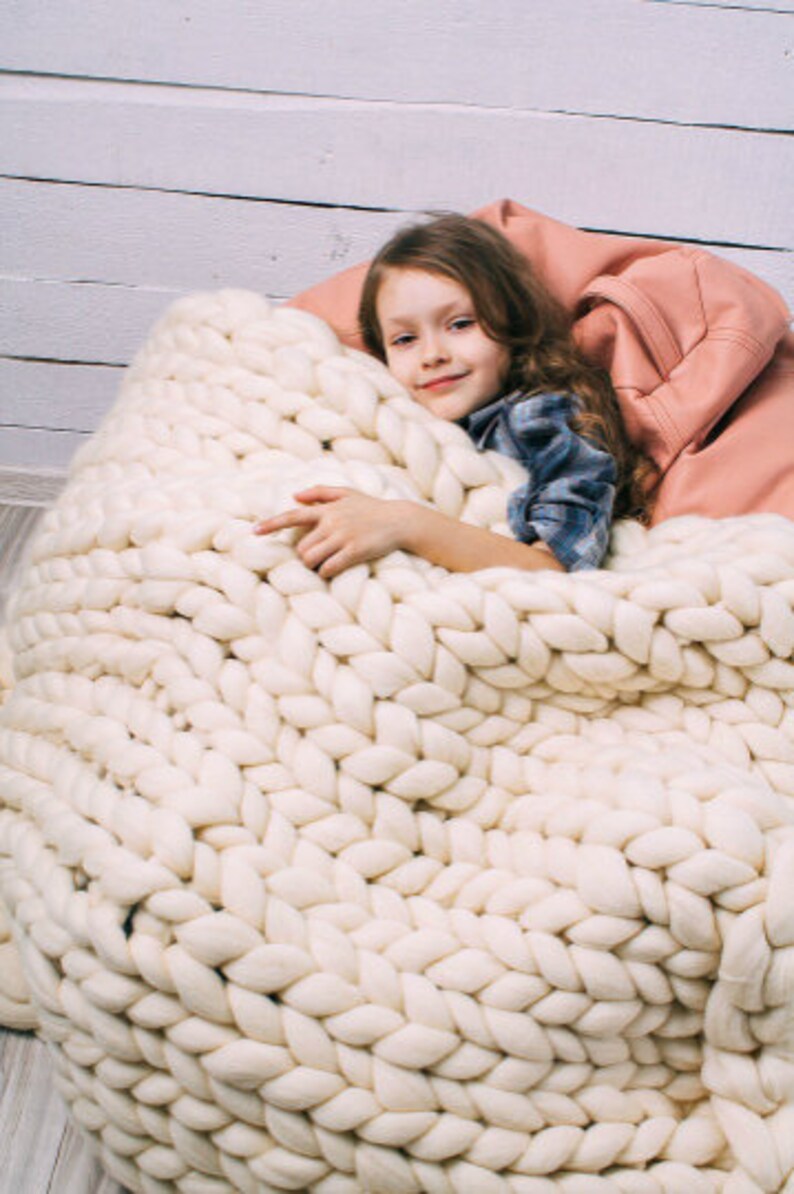 May include: A young person is curled up in a large, white, chunky knit blanket. The blanket is made of thick, soft yarn and has a cozy, comforting look.