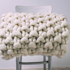 May include: A large, white, chunky knit throw blanket draped over a white chair. The blanket is made of thick yarn and has a soft, fluffy texture.