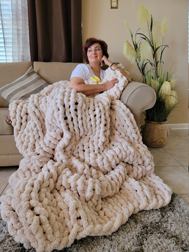 May include: A woman is sitting on a couch with a large, chunky knit blanket in a cream color. The blanket is made of thick yarn and has a soft, fluffy texture.