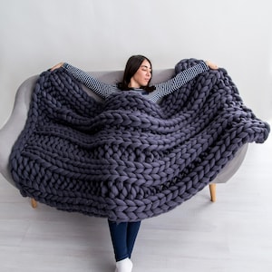 May include: A woman is relaxing on a couch, wrapped in a large, chunky knit gray blanket. The blanket is made of thick yarn and has a loose, textured weave.