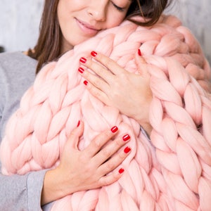 May include: A woman is holding a large, chunky knit blanket. The blanket is a pale pink color and is made of thick yarn. The woman is wearing a gray sweater and has red nail polish on her nails.