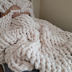 May include: A large, cream-colored chunky knit blanket made with thick yarn. The blanket is draped over a chair and has a soft, fluffy texture.