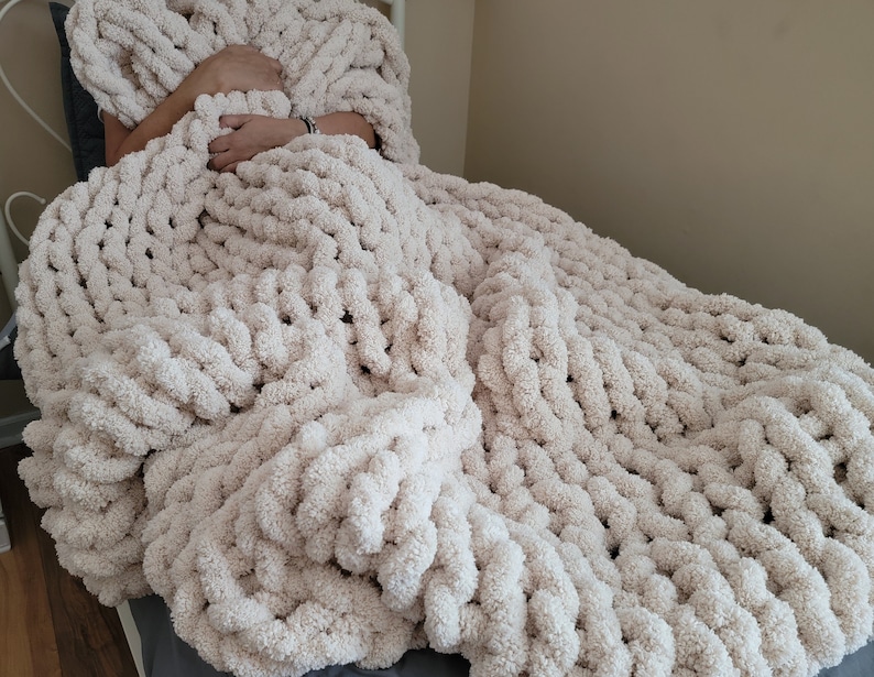 May include: A large, cream-colored chunky knit blanket made with thick yarn. The blanket is draped over a chair and has a soft, fluffy texture.