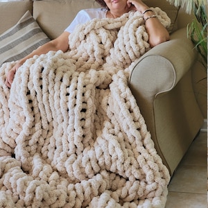 May include: A large, cream-colored chunky knit blanket is draped over a couch. The blanket is made of thick, soft yarn and has a textured, woven pattern.